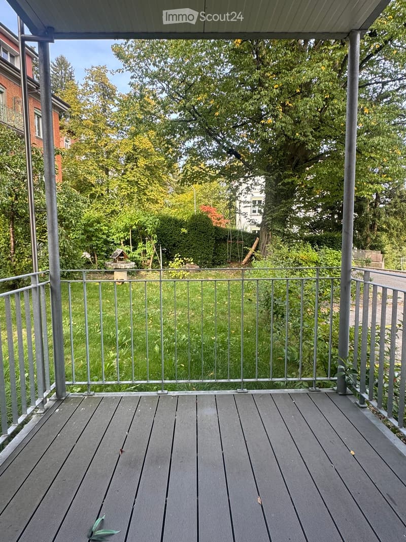 Wooden balcony with metal railing, overlooking a green garden area with trees and shrubs.