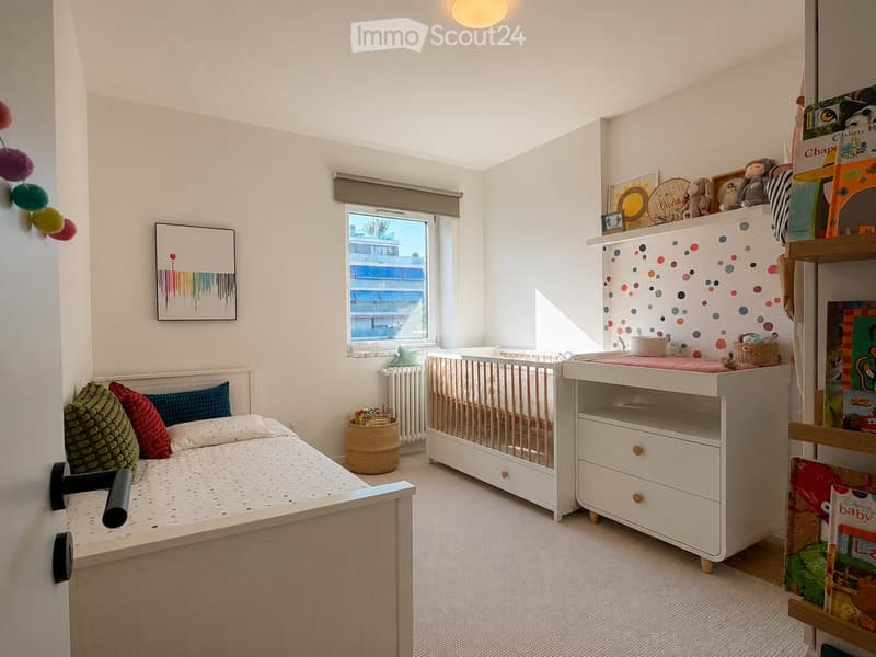 Bright, cozy children's bedroom with a single bed, a crib, a changing table, shelves filled with toys and books, and a window offering a view of the building outside.