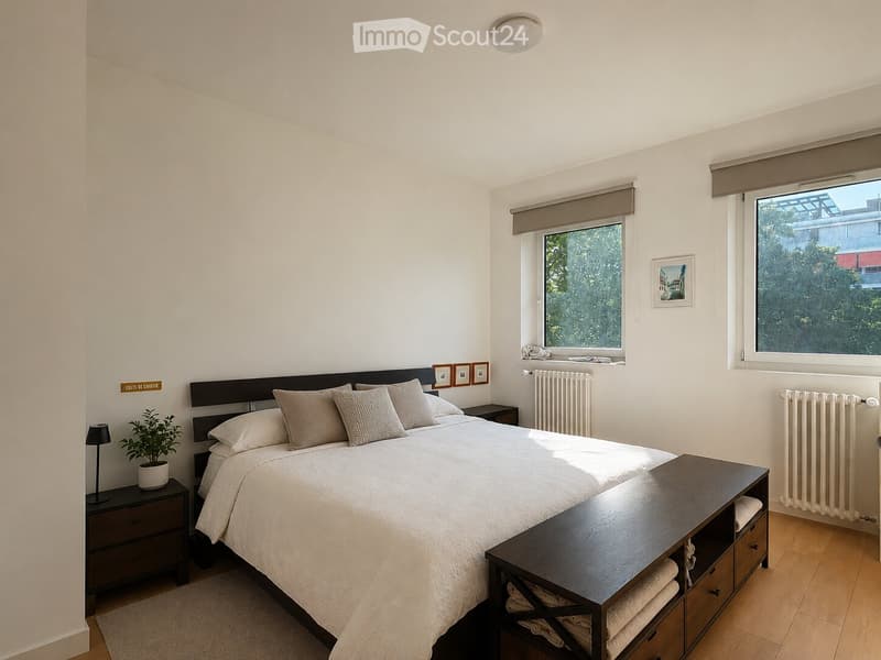A well-lit bedroom with a large bed, nightstands, a potted plant, and a dresser. The room features wooden flooring, white walls, and large windows with blinds.
