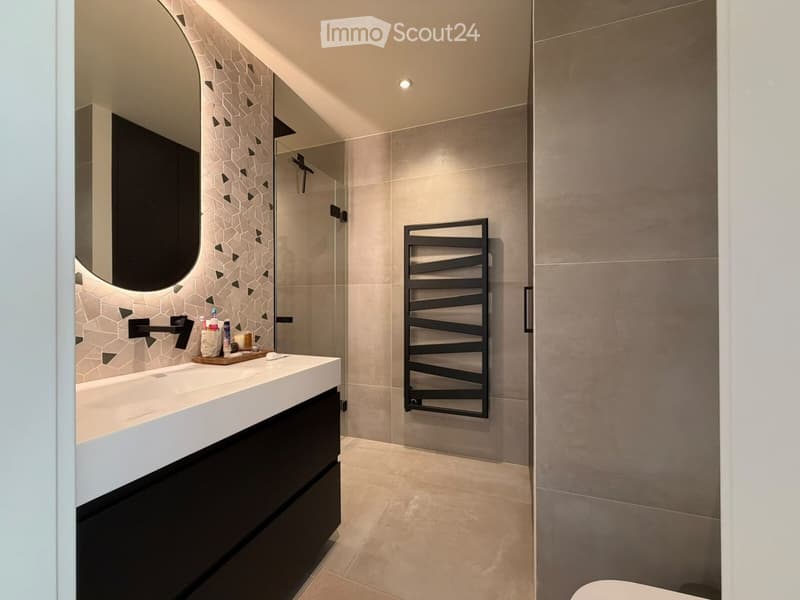Modern bathroom with a white countertop sink, black faucet, large round mirror, and a towel rack in the shower area.