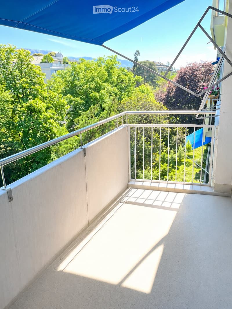 Balcony with metal railing, concrete floor, and a blue awning, overlooking a green, tree-filled area with distant mountains.