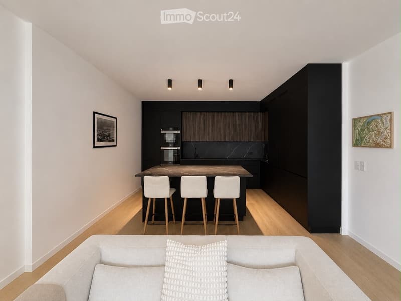 Modern living and dining area with a grey sofa, kitchen island, three white stools, and sleek black kitchen cabinets.