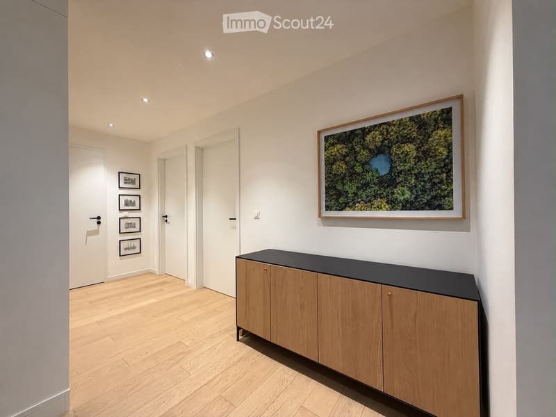 Modern hallway with light wooden flooring, white walls, recessed ceiling lights, and a sleek wooden console table with a black top. Several framed artworks are displayed on the walls, and multiple doors lead to other rooms.