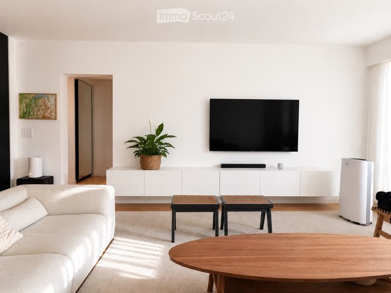 Modern living room with white walls, light wood flooring, a white sofa, a wooden coffee table, two small stools, a potted plant, a flat-screen TV mounted on the wall, and a white cabinet with a lamp on top.