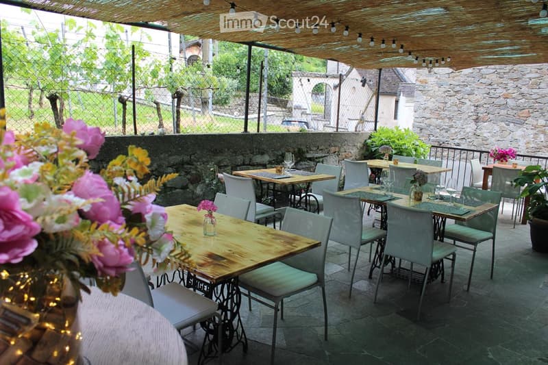 An outdoor dining area with wooden tables, white chairs, and string lights under a thatched roof, surrounded by greenery and stone walls.