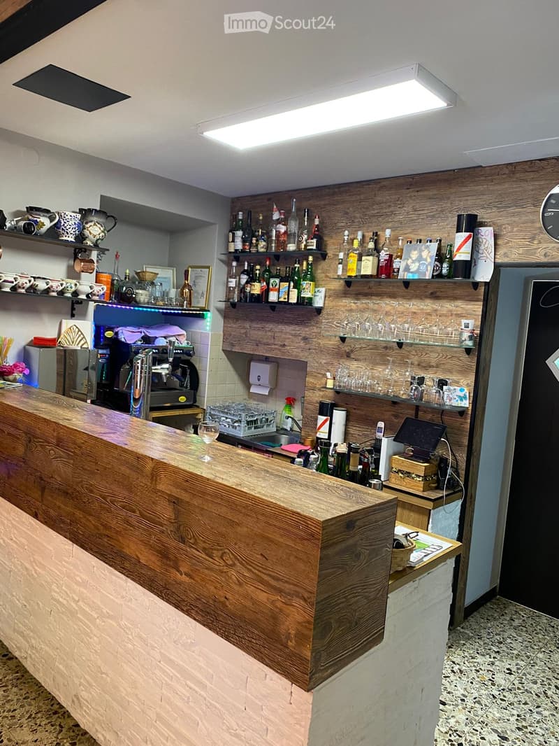 A modern bar area with a wooden counter, shelves filled with wine bottles and glasses, a coffee machine, and a small table with a chair.
