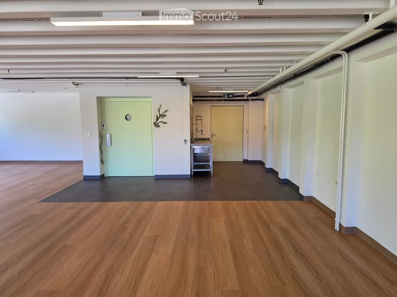 Empty hallway with wooden flooring, white walls, and exposed ceiling pipes. Features a green door, a small cart, and a decorative leaf design on one wall.