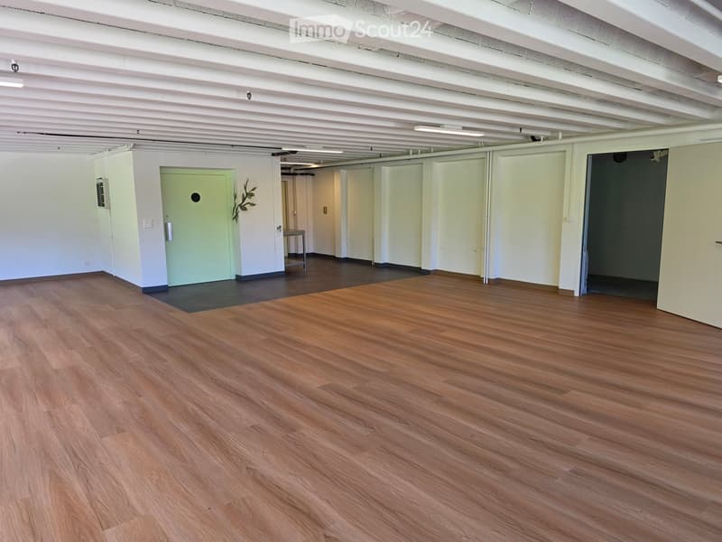 Empty room with wooden flooring, white walls, and several doors leading to other spaces. The room is well-lit with exposed ceiling beams.