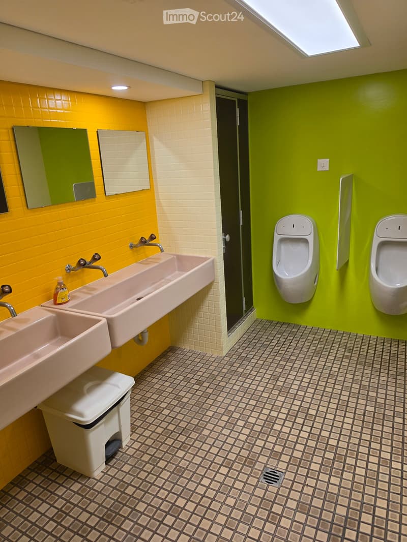 Brightly colored bathroom with yellow and green walls, featuring two sinks, two urinals, mirrors, and a toilet. The floor has a patterned tile design.