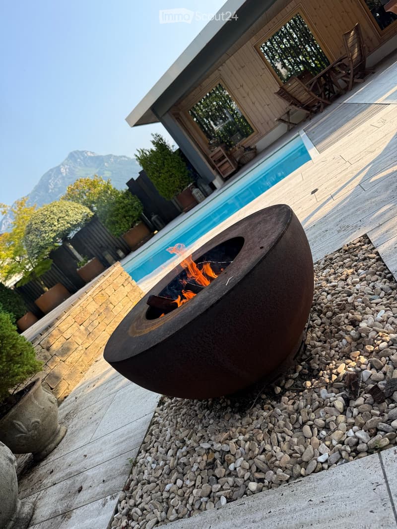 Modern outdoor area with a round metal fire pit surrounded by pebbles, a swimming pool, and a view of mountains.