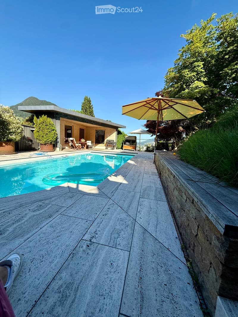 Outdoor area with a swimming pool, patio, and surrounding greenery, featuring a modern house in the background and a scenic mountain view.