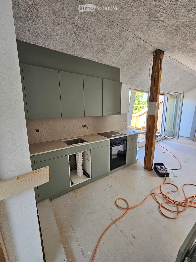 Modern kitchen with grey cabinets, countertop, sink, and oven. Exposed wooden beam and open floor plan with visible construction materials and tools.