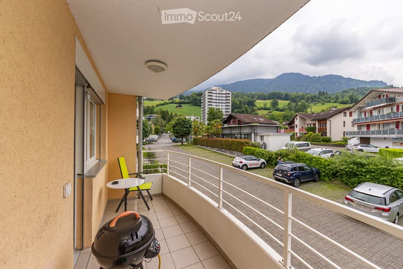 Balcony with a view of a residential area and mountains, equipped with a table, chairs, and a grill.