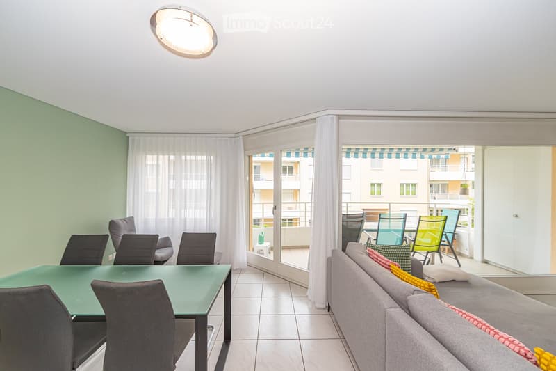 A modern living room with a grey sofa, a glass dining table, and chairs, leading to a balcony with outdoor seating.