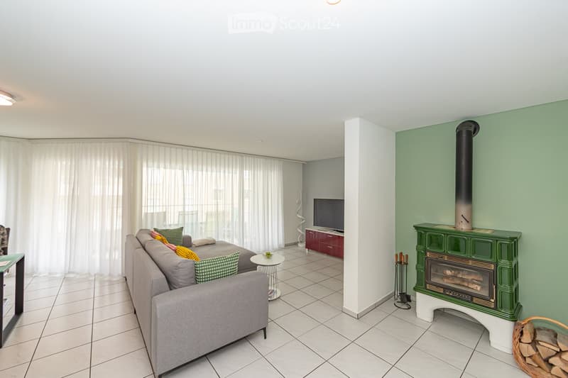 Modern living room with a grey sofa, coffee table, television, and a green fireplace with a chimney. The room features large windows with white curtains and a tiled floor.