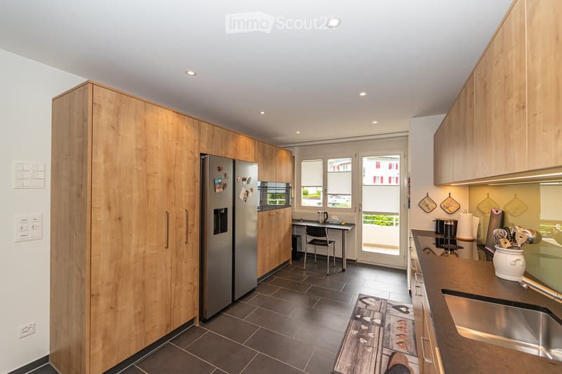 Modern kitchen with wooden cabinets, stainless steel appliances, and a dark tiled floor. Features include a refrigerator, sink, counter, chair, table, and various kitchen utensils.