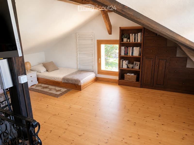 Cozy attic bedroom with wooden floors, slanted wooden ceiling beams, a single bed with pillows and a blanket, a nightstand with a lamp, a built-in bookshelf with books and decorative items, a wooden cabinet, a radiator, and a small rug.