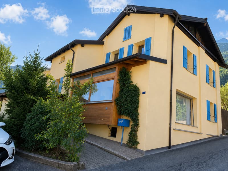 Two-story yellow house with blue shutters, wooden front door, and ivy-covered wall. The house features a balcony and is surrounded by greenery and a parked car.