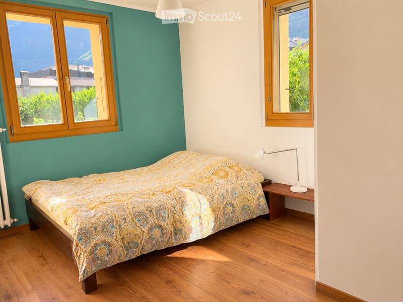 Cozy bedroom with wooden flooring, a large bed with patterned bedding, a nightstand with a lamp, and large windows with wooden frames offering a view of greenery.