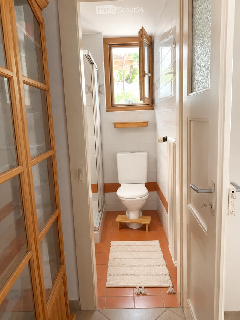 Bathroom with white toilet, wooden stool, white rug, tiled floor, and window with wooden frame.