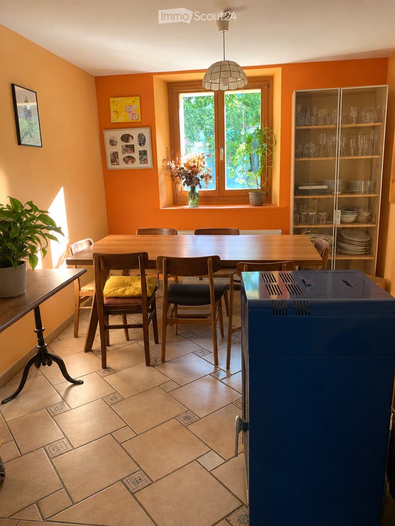 Bright dining area with a wooden table, chairs, a glass cabinet, and potted plants. The room features warm orange walls, tiled flooring, and large windows with a view of greenery.
