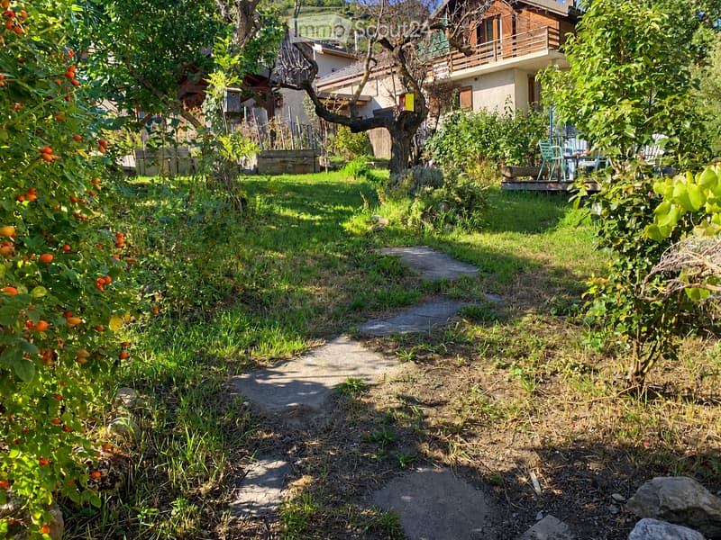 A lush garden with a pathway leading to a house, featuring fruit trees, grass, and outdoor seating on a wooden deck.
