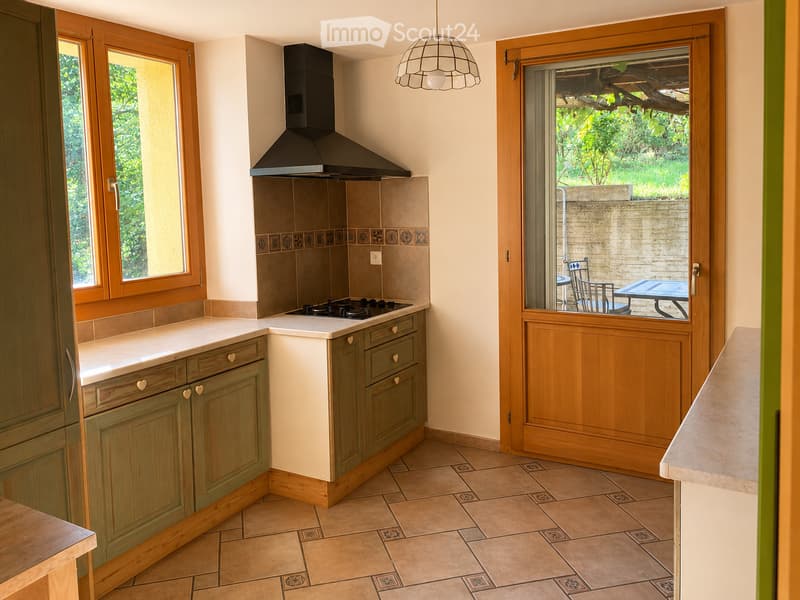 Modern kitchen with light green cabinets, white countertops, a central island, and a large window overlooking a garden. A sliding glass door leads to an outdoor patio with a table and chairs.
