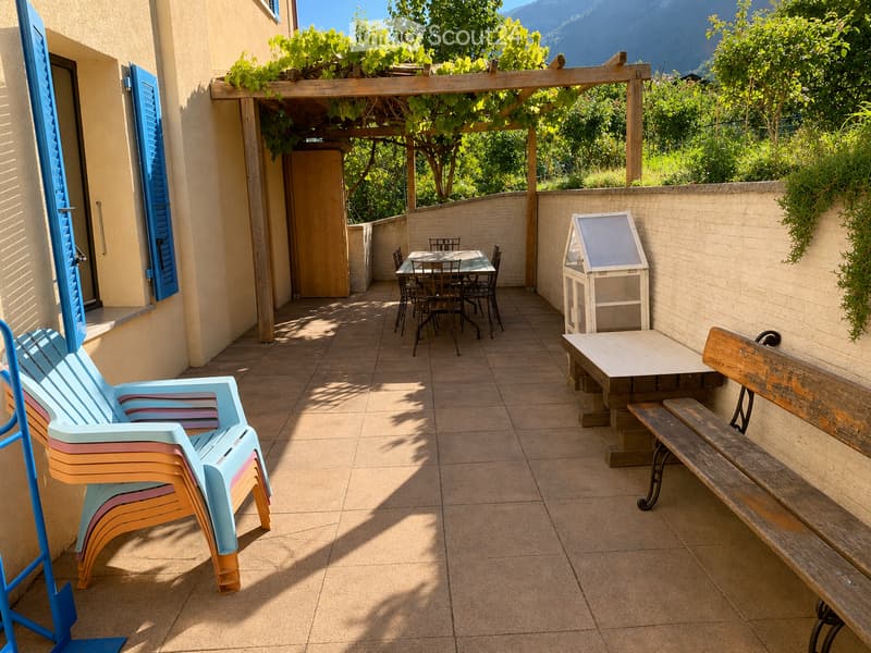 A sunny outdoor patio with a tiled floor, a wooden pergola, colorful plastic chairs, a wooden bench, a small table, and a dining set with chairs.