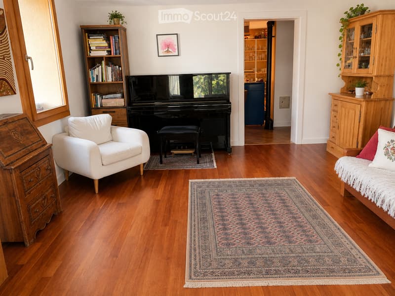 A well-furnished living room with hardwood flooring, a large area rug, a white leather armchair, a wooden bookshelf, a grand piano, and a small table with a plant on it.