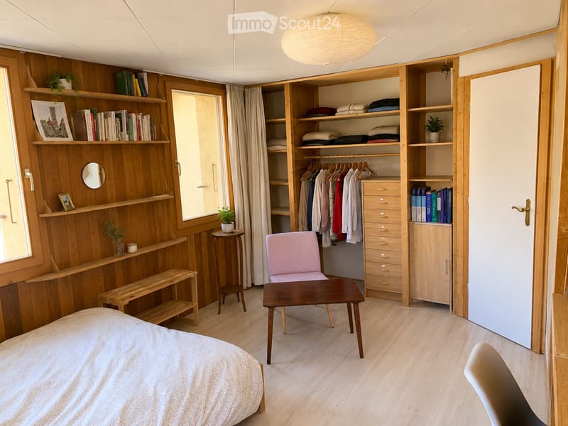 Cozy bedroom with wooden walls and flooring, featuring a bed, a small table, a pink chair, a bench, a built-in wardrobe with hanging clothes, and shelves with books and plants.