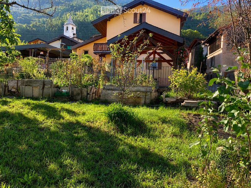 A well-maintained garden with a grassy lawn, wooden planters, and a small wooden fence. The background features a yellow house with a balcony and a small chapel in the distance.