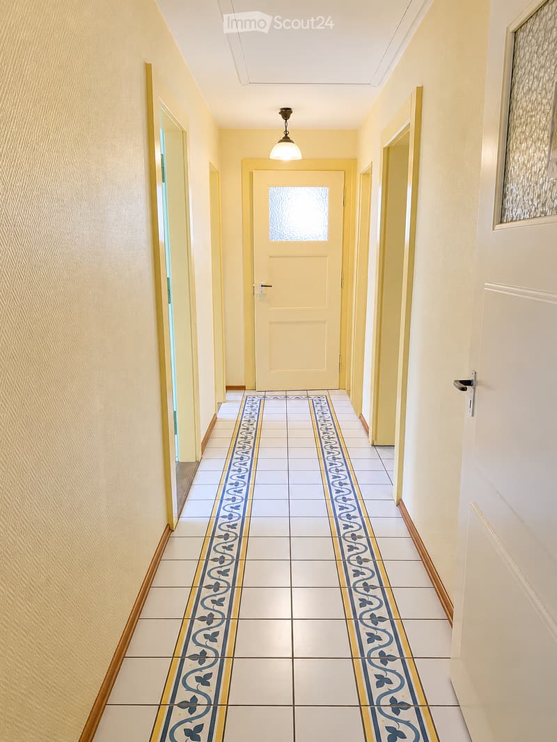 A long hallway with light yellow walls, white ceiling, and decorative blue and gold patterned tiles on the floor. A single pendant light hangs from the ceiling, and several doors line the walls.