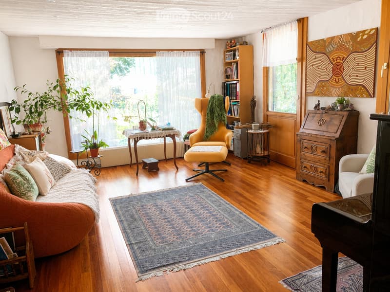 Cozy living room with wooden floors, large windows, a patterned rug, a sofa, a yellow armchair, a small table, a bookshelf, and a piano.