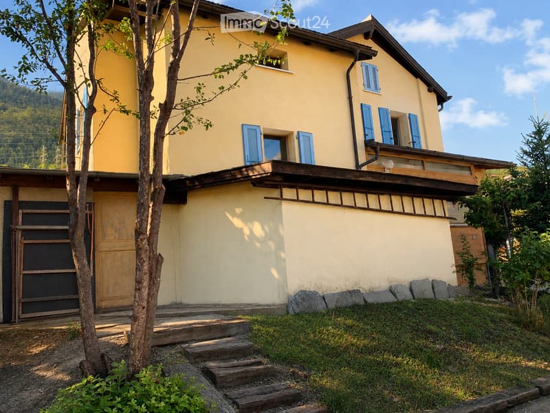 Two-story yellow house with blue shutters, wooden balcony, stone steps, and surrounding greenery.
