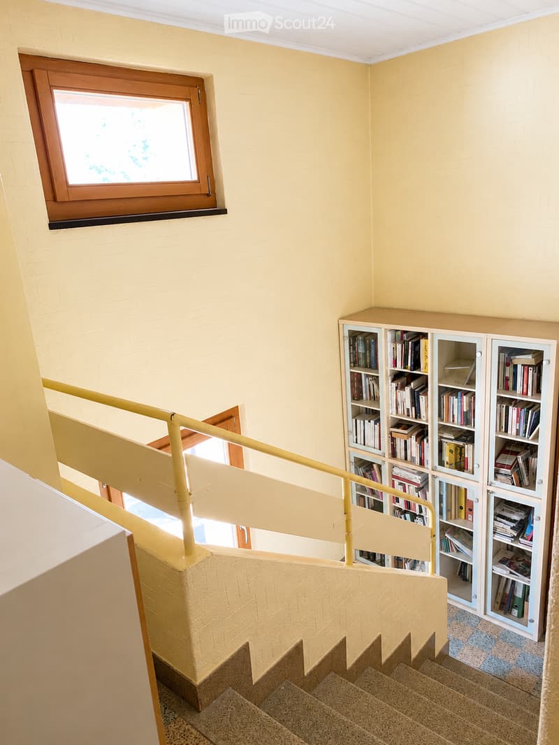 Staircase with yellow railing, light yellow walls, and a window overlooking the outside. A bookshelf filled with books is visible on the right side.