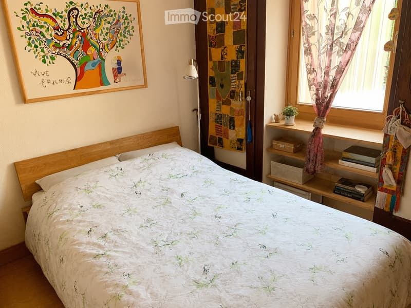 Cozy bedroom with a wooden bed frame, floral bedding, a nightstand with a lamp, a shelf with books and a small plant, and a colorful patchwork tapestry on the wall.