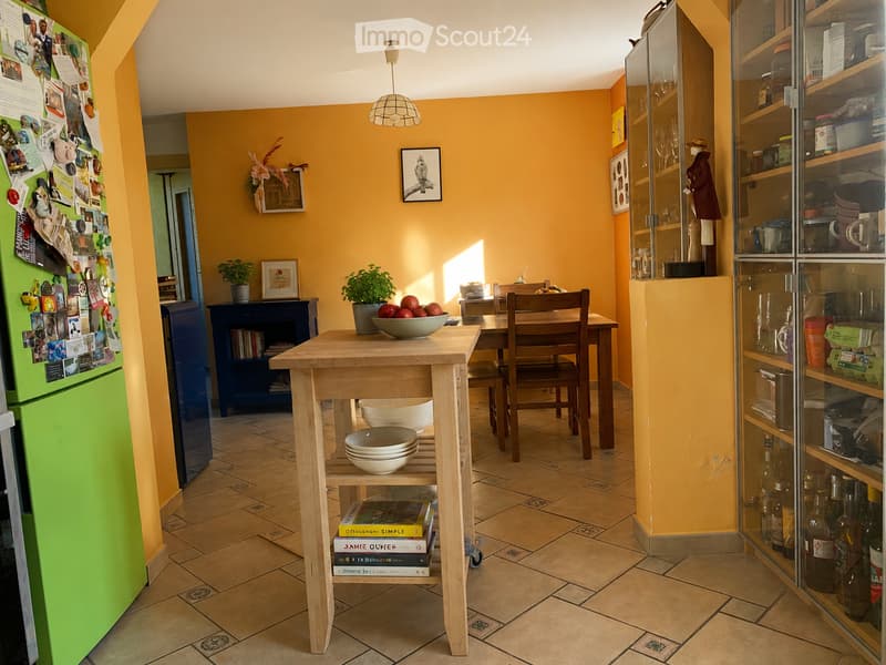 Bright kitchen with yellow walls, tiled floor, and natural light. Features a green refrigerator, wooden table with bowls, chairs, glass cabinet, and potted plants.