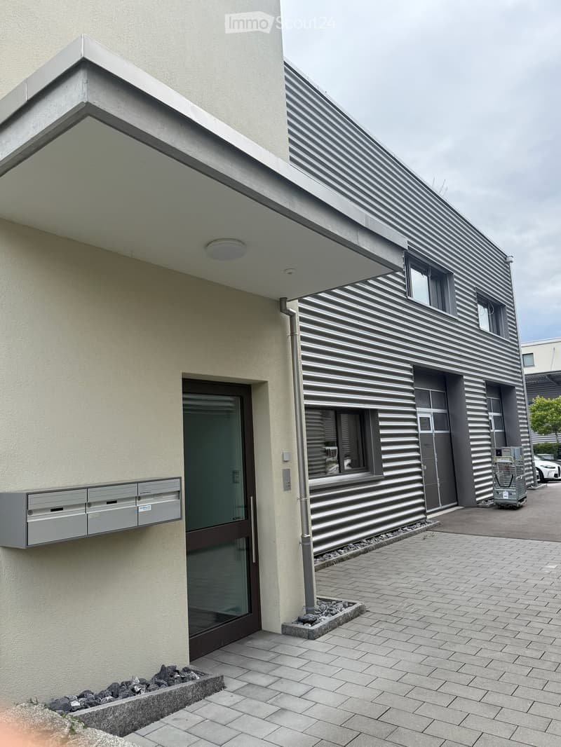 Modern building entrance with a glass door, mailboxes, and a textured facade. The entrance features a raised platform with stone borders and a paved walkway.