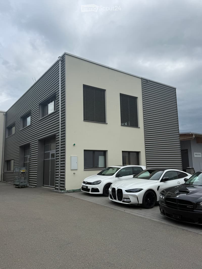 Modern two-story building with a combination of white and dark grey exterior walls, multiple windows, and a large entrance door. Several cars are parked in front of the building.