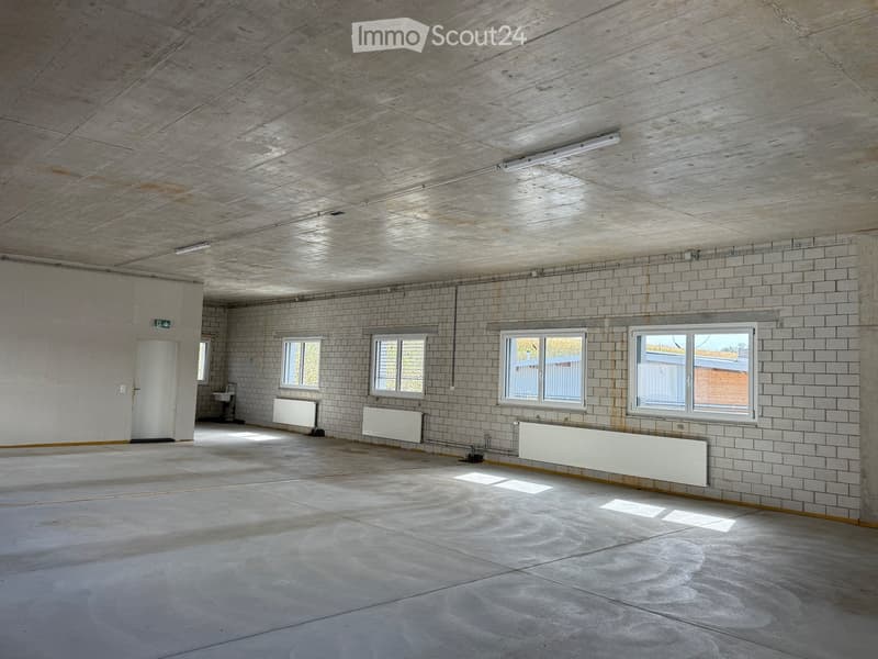 Empty room with concrete ceiling, tiled walls, and large windows. Features include a door, radiators, and a sink in the background.