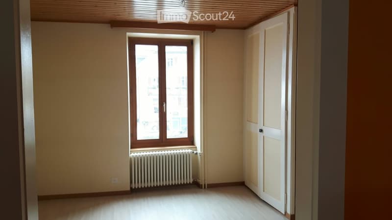 Empty room with wooden flooring, white walls, a window with wooden frame, radiator, and a closed door.