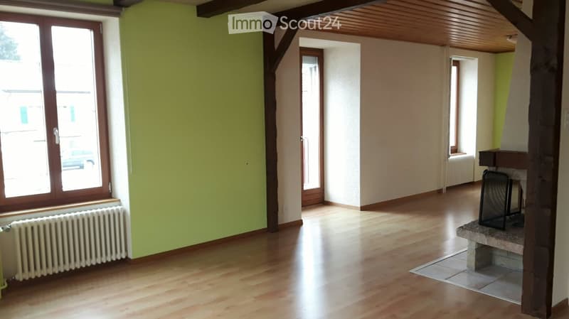 Empty room with wooden flooring, exposed wooden beams, large windows, radiator, and fireplace area.