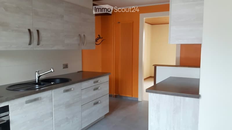 Modern kitchen with white cabinets, dark countertops, and a stainless steel sink. The space features a bright orange accent wall and a visible doorway leading to another room.