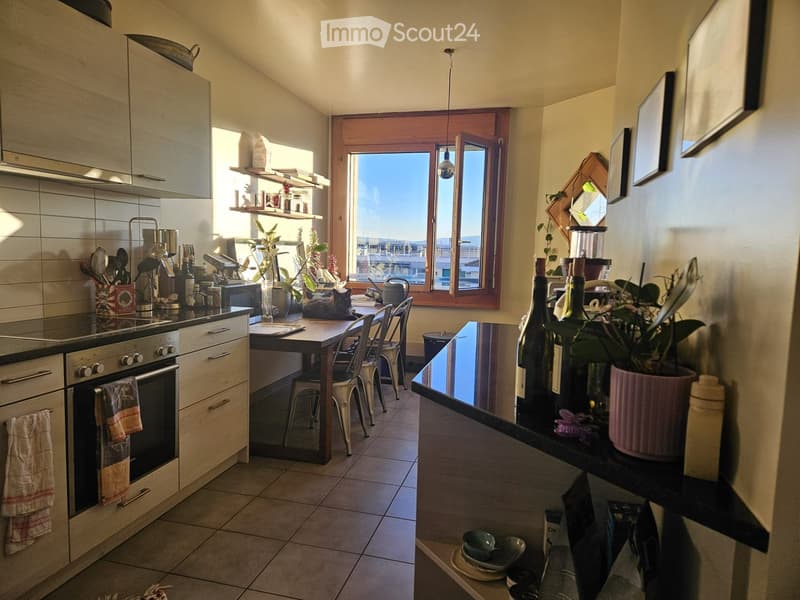 Bright kitchen with wooden cabinets, tiled floor, and a large window offering a view. Includes an oven, table with chairs, a blender, and various kitchen items.