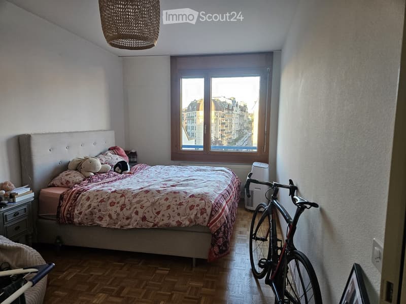 Cozy bedroom with a large window overlooking a cityscape, featuring a bed with floral bedding, a nightstand with books, a bicycle, and a wicker hanging lamp.