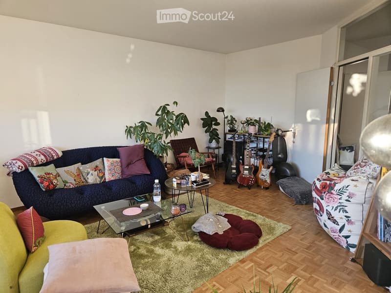 A cozy living room with a dark blue sofa, colorful pillows, a glass coffee table, a yellow armchair, a floral-patterned chair, a guitar stand with guitars, a large plant, and a round balloon.