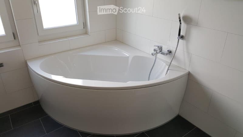 Modern bathroom with a corner bathtub, white tiles, and a window above the tub.