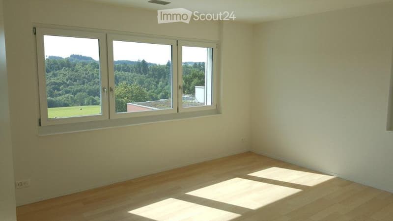 Empty room with light wooden flooring and large windows offering a view of greenery outside.