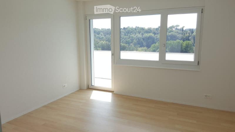 Empty room with wooden flooring, large windows, and a sliding glass door leading to a balcony with a scenic view of trees.