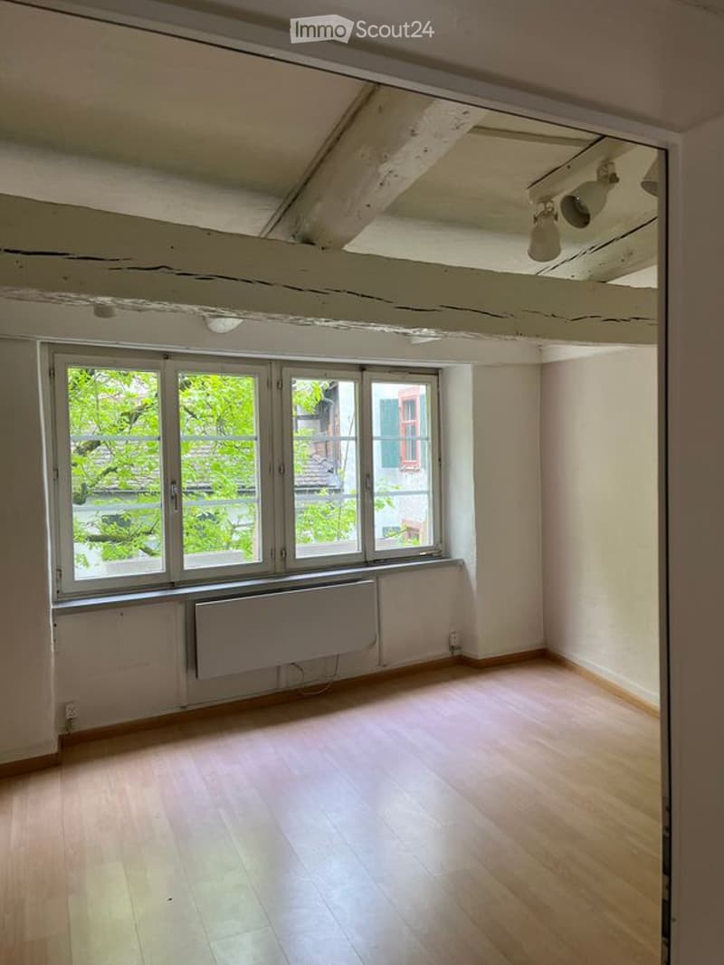 Empty room with wooden flooring, large windows with white frames, radiator, and exposed ceiling beams.